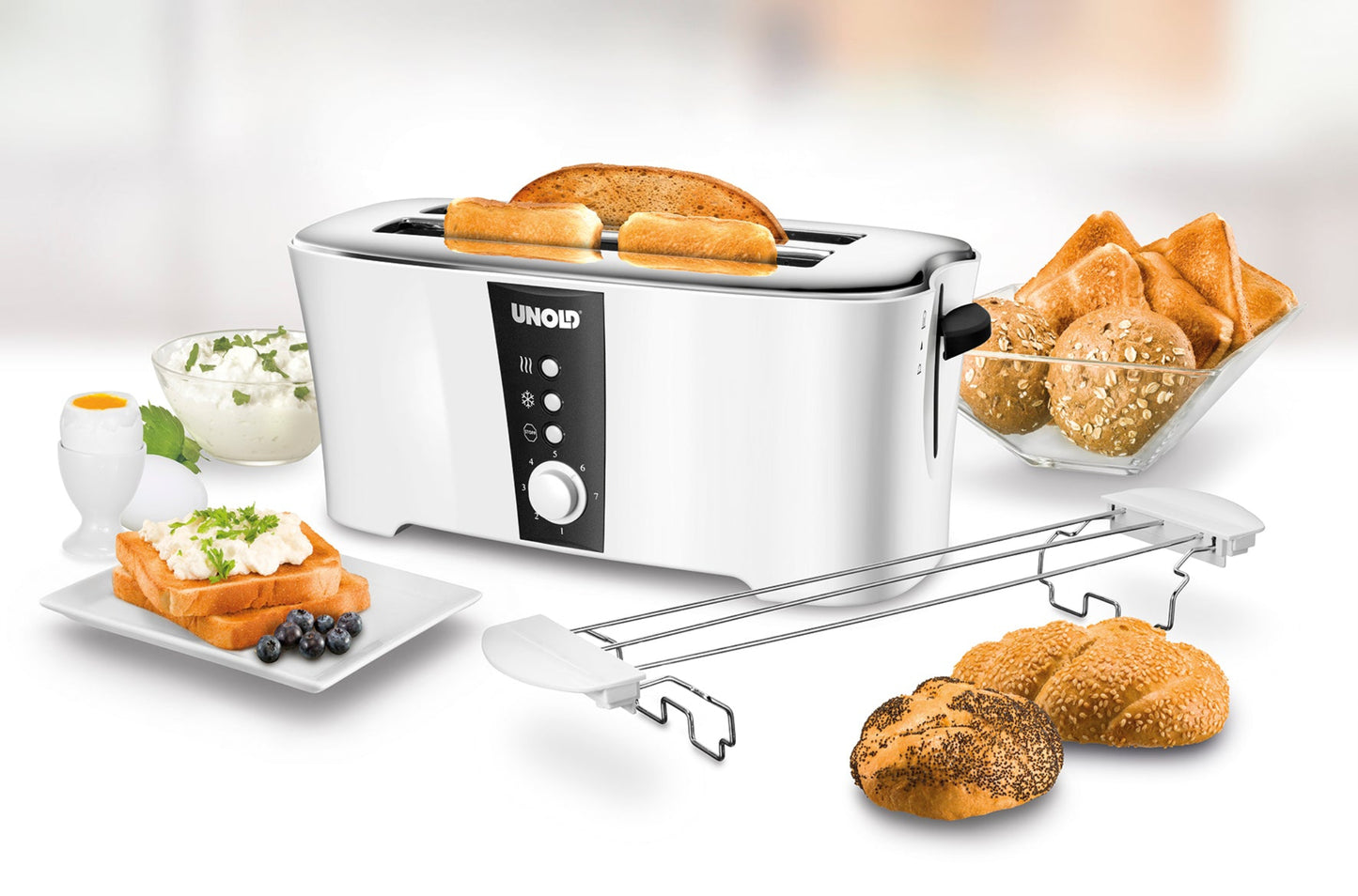 Der Unold Toaster Design Dual in Weiß toastet Brot auf einer weißen Fläche, umgeben von verschiedenen Brötchen, einem Teller Toast, Blaubeeren, einem weichgekochten Ei und Hüttenkäse.