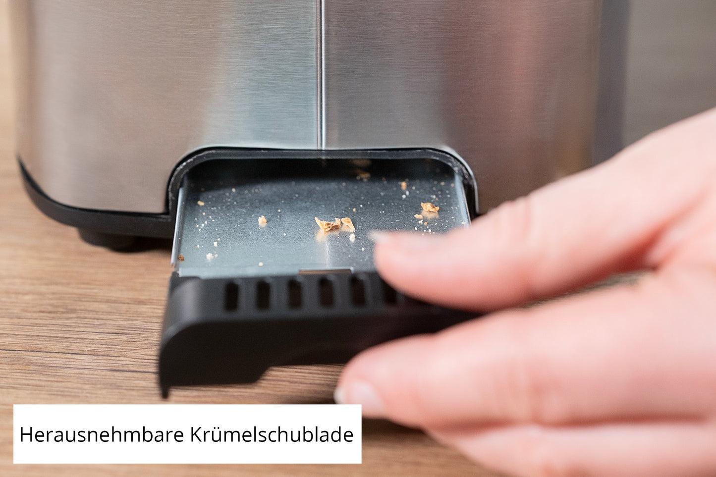 Eine Hand entfernt die Krümelschublade aus dem Unold Toaster Tom und gibt darin einige Krümel frei. Der deutsche Ausdruck „Herausnehmbare Krümelschublade“ bedeutet „herausnehmbare Krümelschublade“.