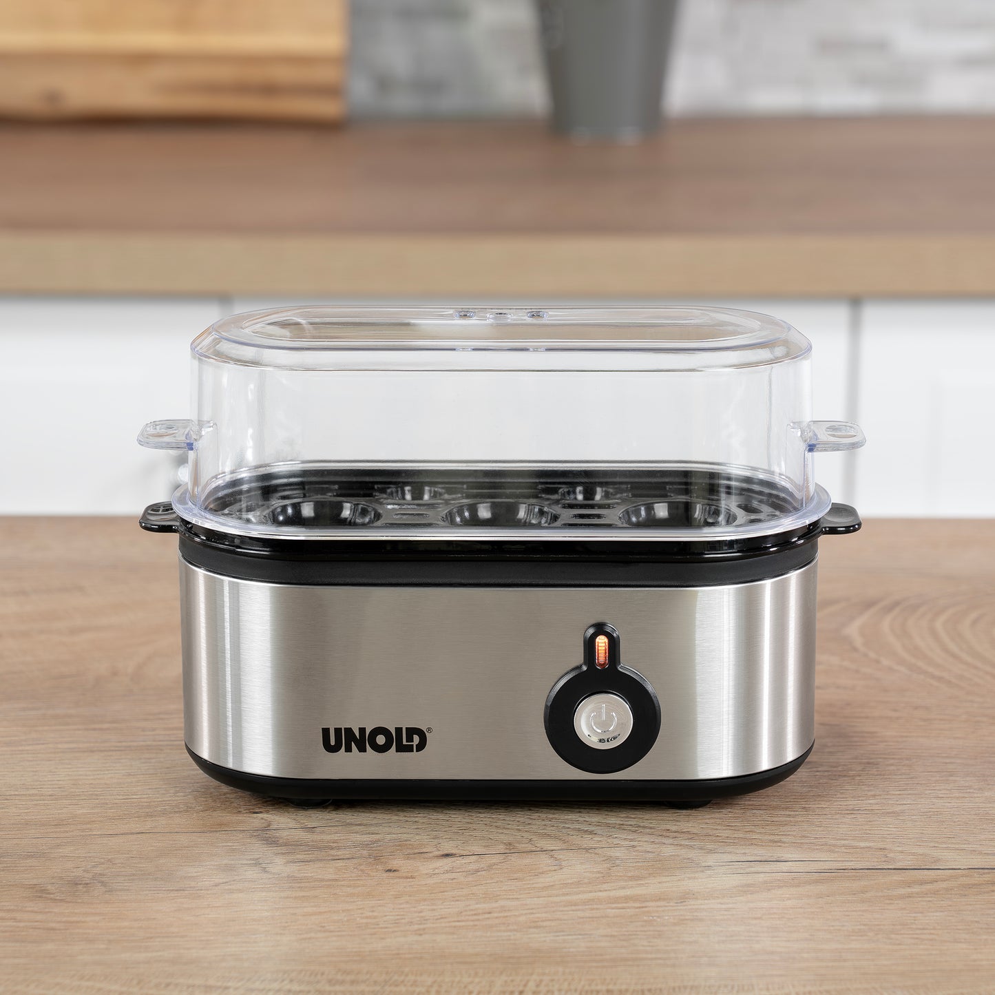 Der Unold Eierkocher Mini, ein Edelstahl-Eierkocher mit einem durchsichtigen Kunststoffdeckel und einem orangefarbenen Einschaltknopf, steht auf einer hölzernen Küchenzeile.