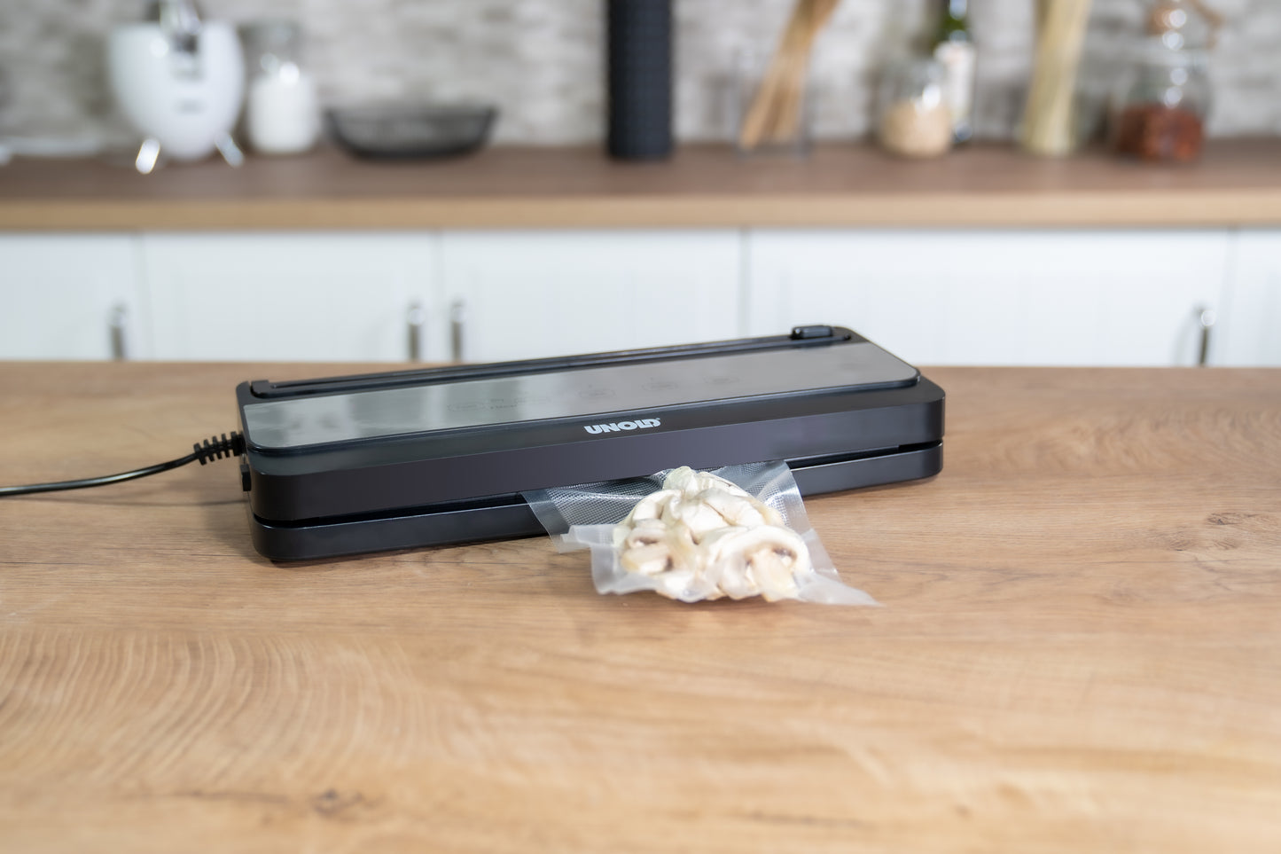 Der Unold Vakuumierer Food Saver Black steht auf einer hölzernen Küchentheke und versiegelt einen Plastikbeutel mit weißen Champignons. Im Hintergrund sind Gläser, Utensilien und Küchengeräte zu sehen.