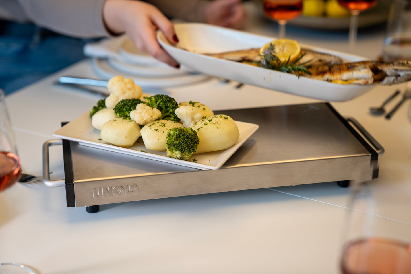Eine Nahaufnahme zeigt die Unold Wärmespeicherplatte Elena, die eine Servierplatte mit Salzkartoffeln, Brokkoli und Blumenkohl warm hält, während eine Person eine Platte mit gegrilltem Fisch mit Zitrone und Kräutern hinzufügt. Gläser mit Roséwein stehen ebenfalls auf dem Tisch.