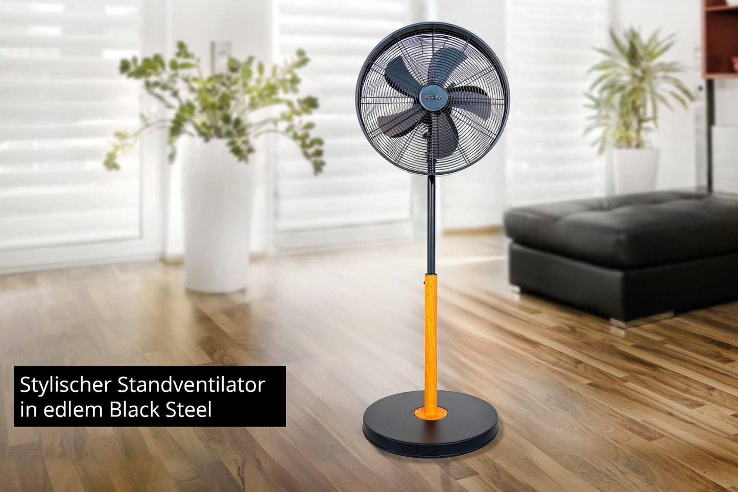 Ein Unold Standventilator Black Steel in modernem Schwarz und Orange steht in einem hellen Wohnzimmer mit Holzboden, großen Fenstern, einem schwarzen Sofa und grünen Pflanzen. Text: Stylischer Standventilator in edlem Black Steel.