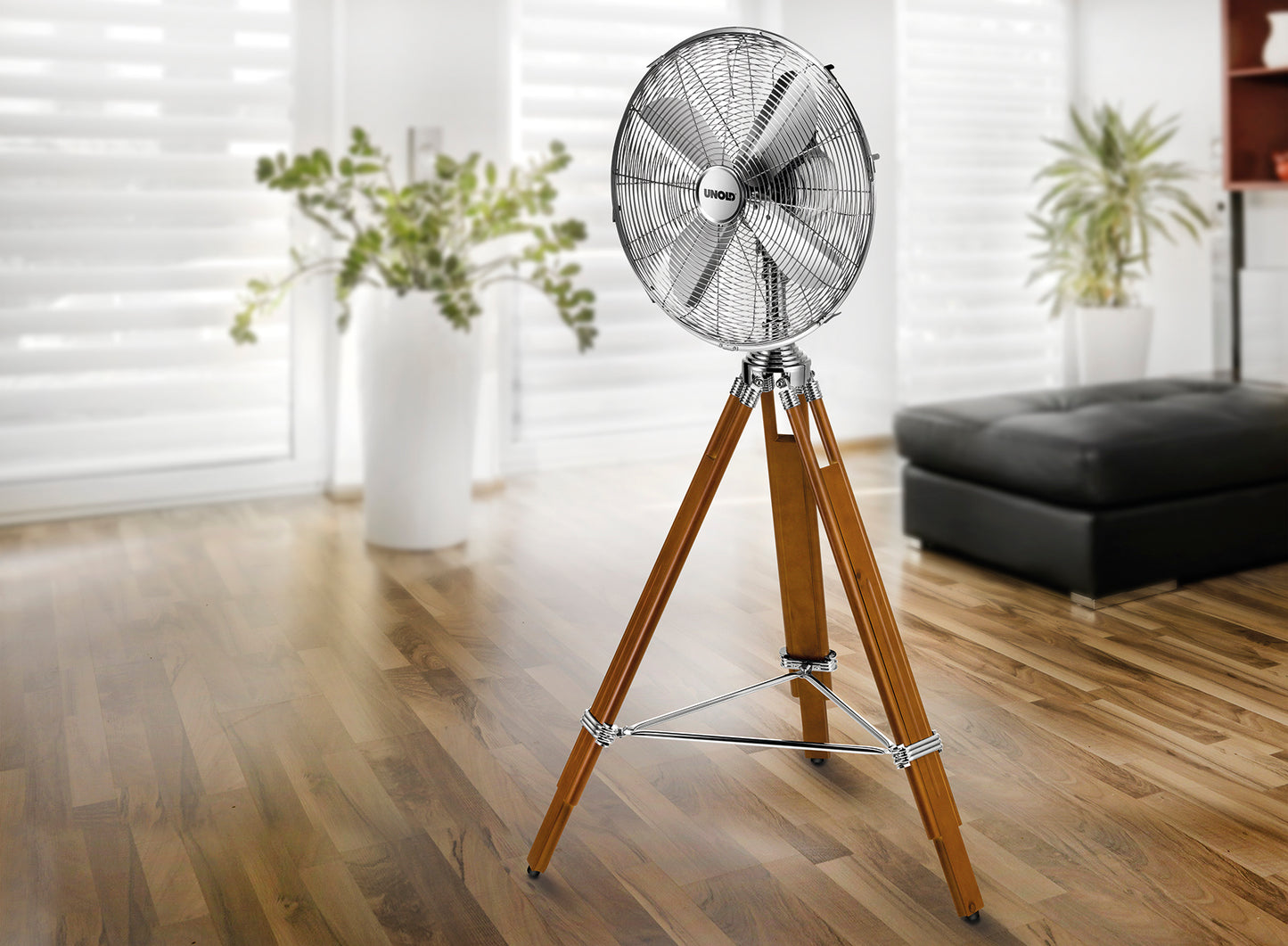 Der Unold STANDVENTILATOR KOLONIAL mit Metallkopf und dreibeinigen Holzbeinen steht auf einem polierten Holzboden in einem hellen Wohnzimmer mit großen Fenstern und Topfpflanzen.
