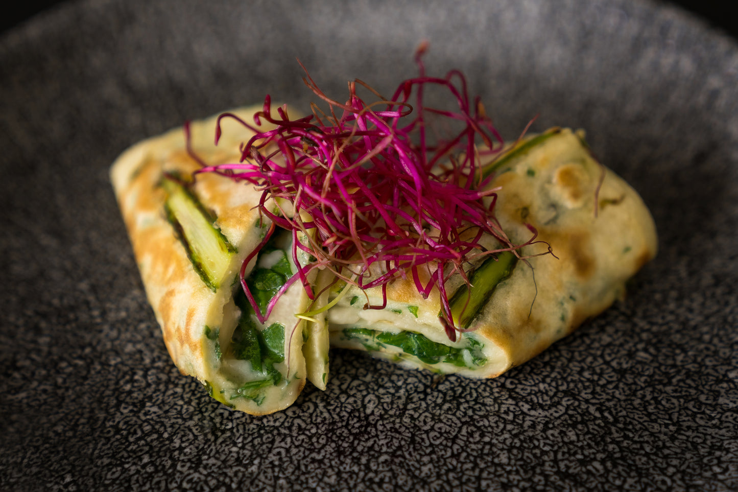 Das Unold Raclette Multi 4 in 1 inklusive Raclette Buch wird auf einem stilvollen dunklen Teller serviert, begleitet von grünem Gemüse und leuchtend violetten Microgreens für eine frische, farbenfrohe Präsentation.