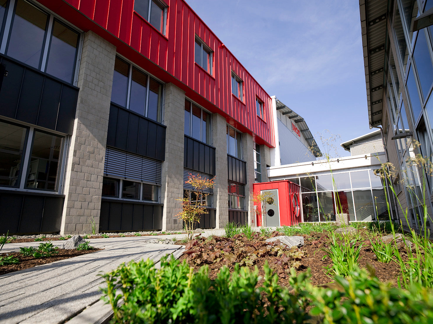 Ein modernes Gebäude mit roten und grauen Außenwänden, großen Fenstern und einem Garten mit jungen Pflanzen und einem Steinweg im Vordergrund unter einem klaren blauen Himmel.