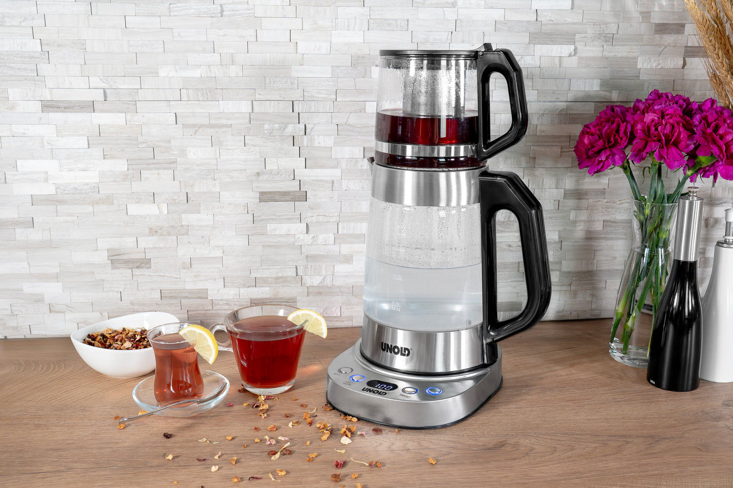 Die Unold AG Samowar Selin, ein moderner elektrischer Teezubereiter mit Glaskannen, brüht Tee auf einer hölzernen Arbeitsplatte neben zwei Tassen mit Zitronenscheiben, losem Tee in einer Schale, verstreuten Blättern und einer Vase mit lila Blumen.