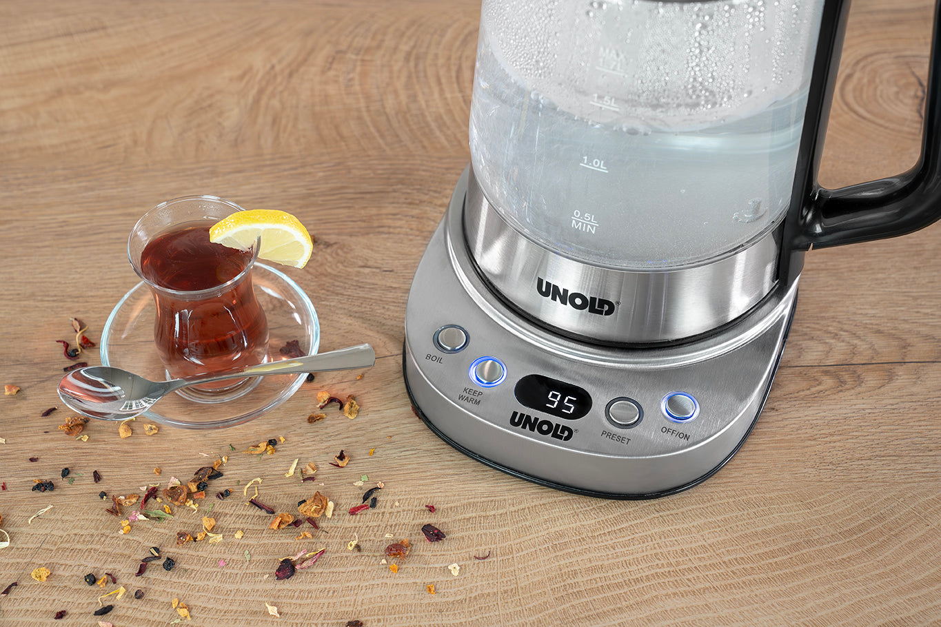 Der gläserne Wasserkocher der Unold AG Samowar Selin mit einer Temperatur von 95°C steht auf einer Holzplatte neben einer Glastasse mit Tee und Zitrone und einem Löffel. Getrocknete Teeblätter liegen in der Nähe verstreut.