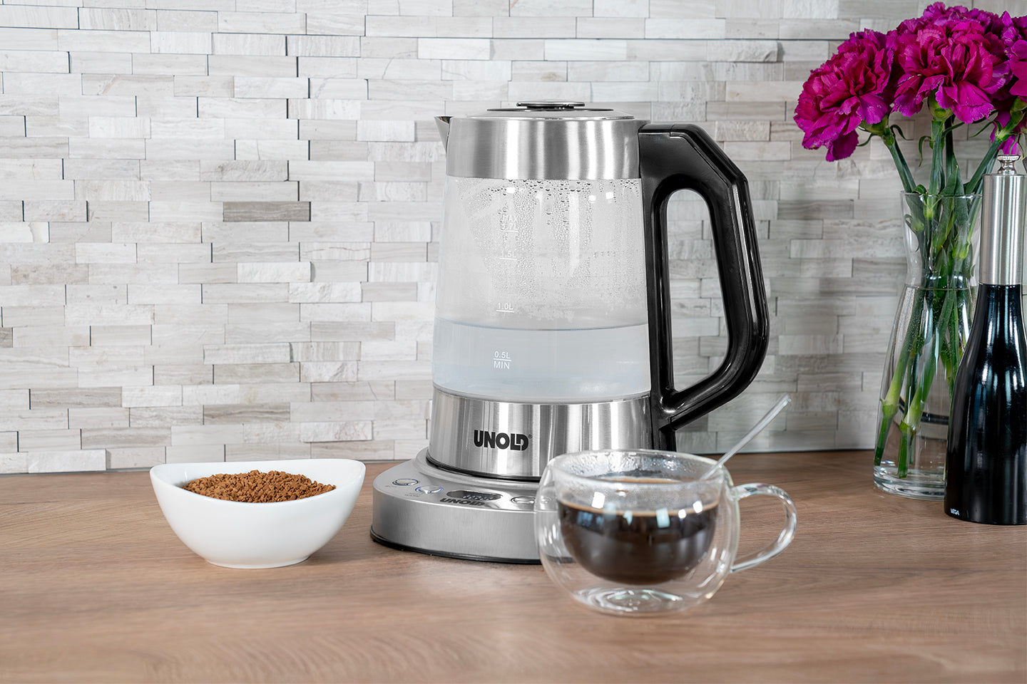 Ein Samowar Selin Wasserkocher von Unold AG steht auf einer hölzernen Arbeitsplatte neben Instantkaffee, einer Glastasse mit Löffel und violetten Blumen, alles vor einer gefliesten Arbeitsplatte.