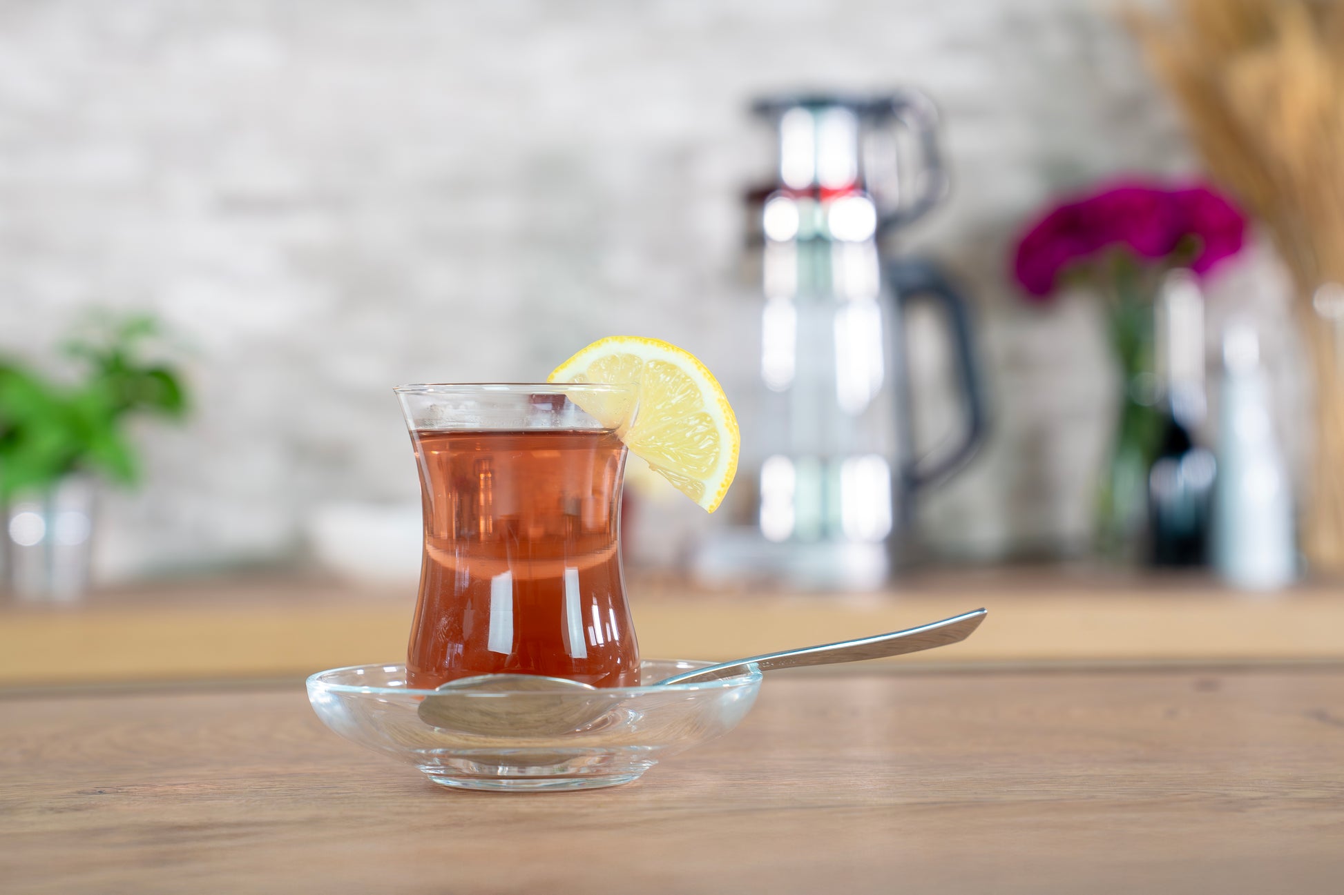 Ein Glas Tee mit einer Zitronenscheibe am Rand, serviert mit einem Löffel auf einer Glasuntertasse, steht auf einem Holztisch. Im unscharfen Hintergrund sind Pflanzen und die Metallteekanne der Unold AG Samowar Selin zu sehen.