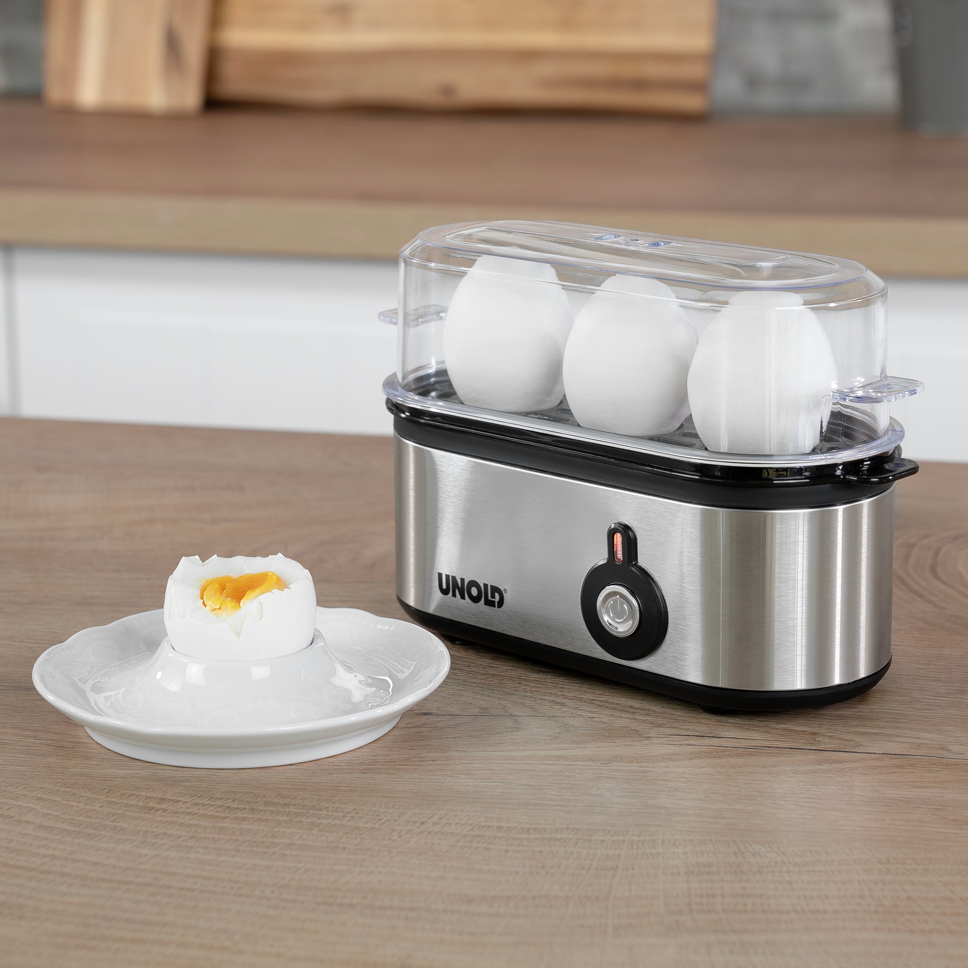 Der Unold Eierkocher Mini mit drei ganzen Eiern steht auf dem Küchentisch. Daneben liegt ein weichgekochtes Ei mit flüssigem Dotter in seiner aufgesprungenen Schale auf einem weißen Teller.