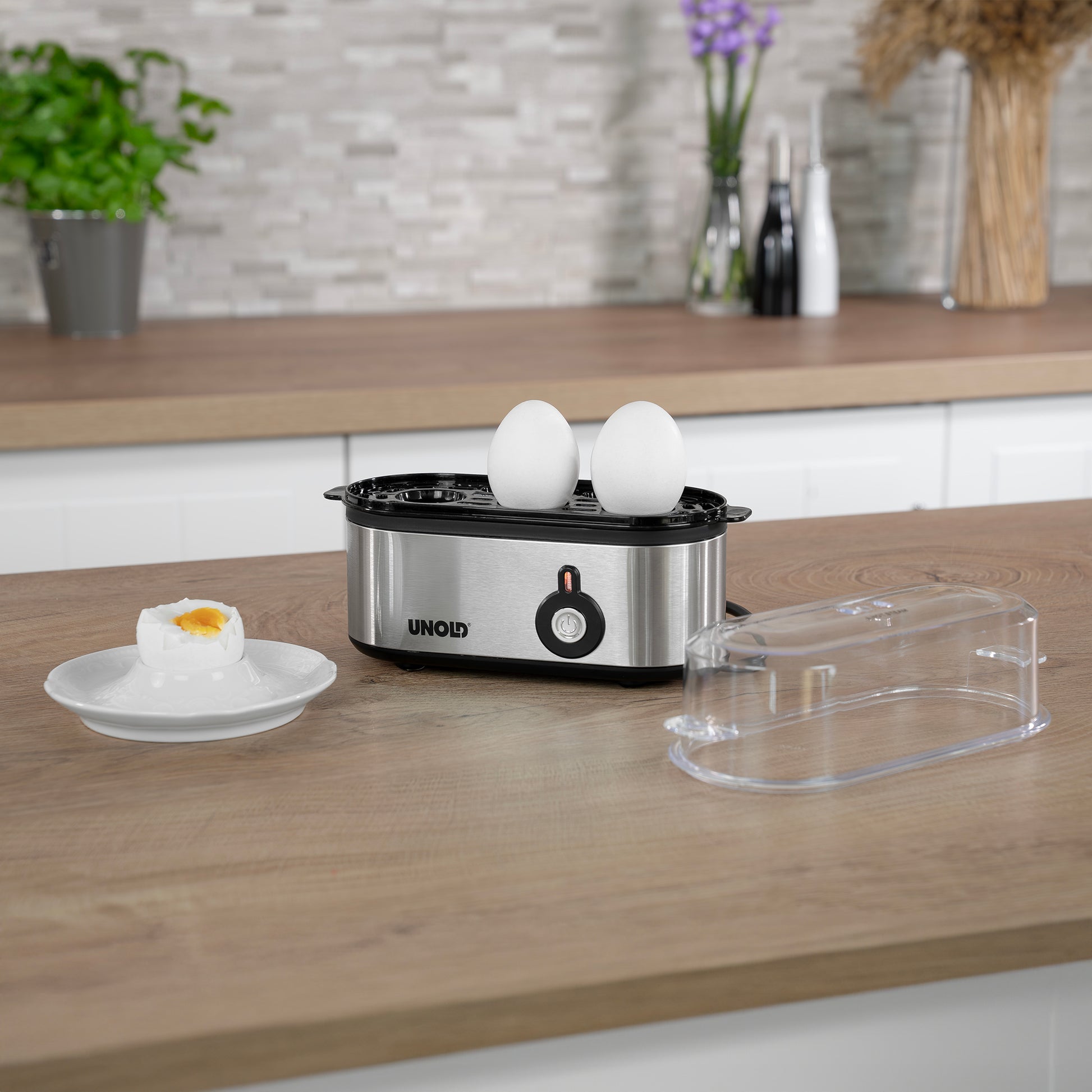 Der Unold Eierkocher Mini mit zwei weißen Eiern steht auf einem Küchentisch neben seinem durchsichtigen Plastikdeckel und einem Teller mit einem weichgekochten Ei. Pflanzen und Utensilien schmücken den Küchenhintergrund.