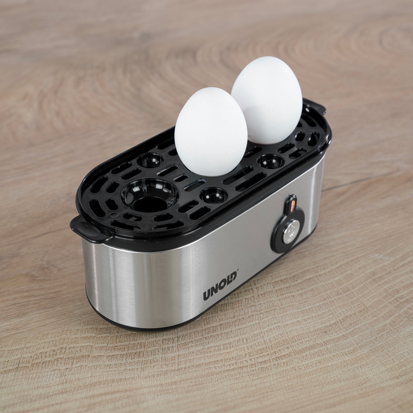 Der Unold Eierkocher Mini, ein elektrischer Eierkocher aus Edelstahl, verfügt über einen schwarzen Kunststoffdeckel mit runden Schlitzen, in die zwei weiße Eier passen, und steht auf einer Holzfläche.