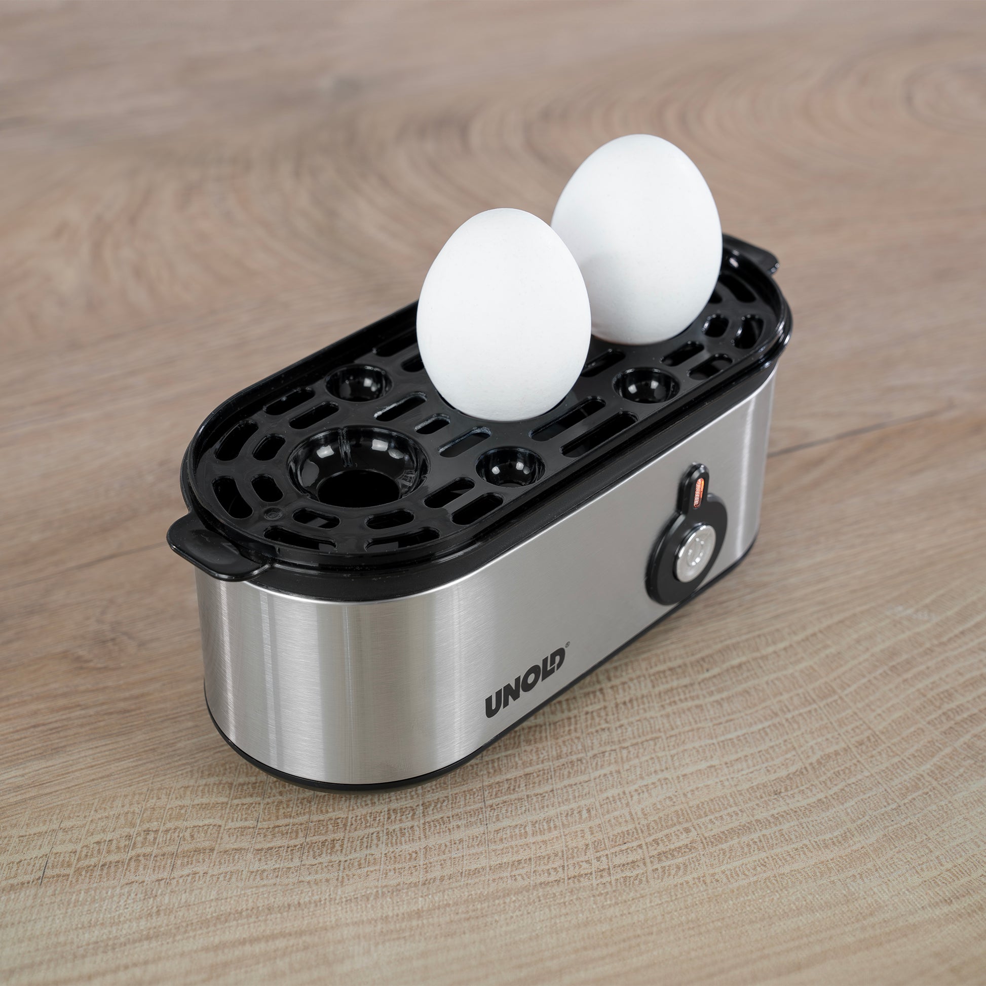 Der Unold Eierkocher Mini, ein elektrischer Eierkocher aus Edelstahl, verfügt über einen schwarzen Kunststoffdeckel mit runden Schlitzen, in die zwei weiße Eier passen, und steht auf einer Holzfläche.