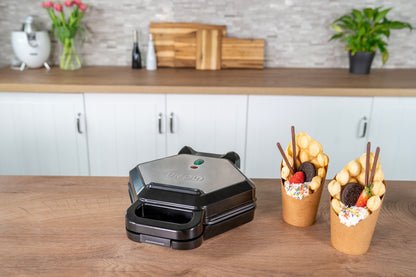 Der Unold Bubble-Waffeleisen Bobby steht auf einer Holztheke. Daneben stehen zwei Bubble-Waffeln mit Früchten, Keksen und Streuseln. Im Hintergrund sind weiße Schränke und Pflanzen zu sehen.