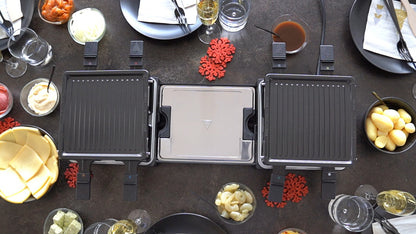 Eine Draufsicht auf den ausziehbaren Raclette-Grill von Unold auf einem dunklen Tisch, umgeben von Tellern, Gläsern, Besteck, kleinen Schüsseln mit Gemüse, Käse, Soßen und festlichen roten Schneeflockendekorationen.