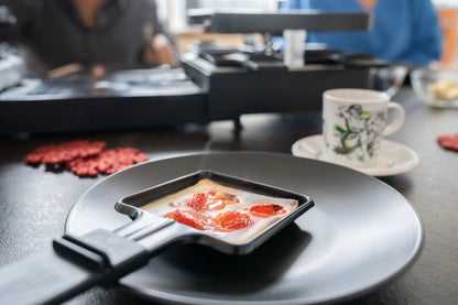 Auf einem schwarzen Teller steht ein ausziehbares Raclette-Pfännchen von Unold mit geschmolzenem Käse und Tomatenscheiben, im Hintergrund sind eine Tasse mit grünem Muster und verschwommene Figuren an einem Tisch zu sehen.