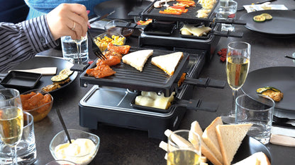 Menschen versammeln sich um einen Tisch und bereiten mit dem ausziehbaren Raclette-Grill von Unold Brot, Gemüse und Käse zu. Getränke, Teller und Besteck stehen auf dem Tisch, während eine Person das Essen auf dem Grill mit einer Gabel wendet.