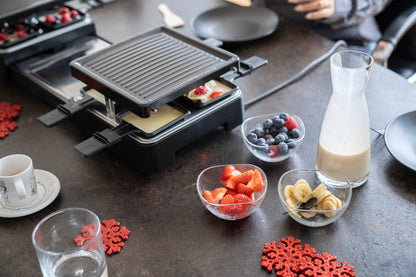 Der ausziehbare Raclette-Grill von Unold schmilzt Käse auf der Tischplatte, umgeben von Schüsseln mit Erdbeeren, Bananen, Blaubeeren, einer Karaffe mit cremiger Flüssigkeit, Tellern, Gläsern und roten Schneeflockendekorationen.