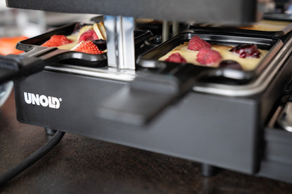 Nahaufnahme eines ausziehbaren Raclette-Grills von Unold auf einer Küchenarbeitsplatte, mit mit Teig gefüllten Schalen und verschiedenen Beeren wie Erdbeeren und Himbeeren, die darin zubereitet werden.