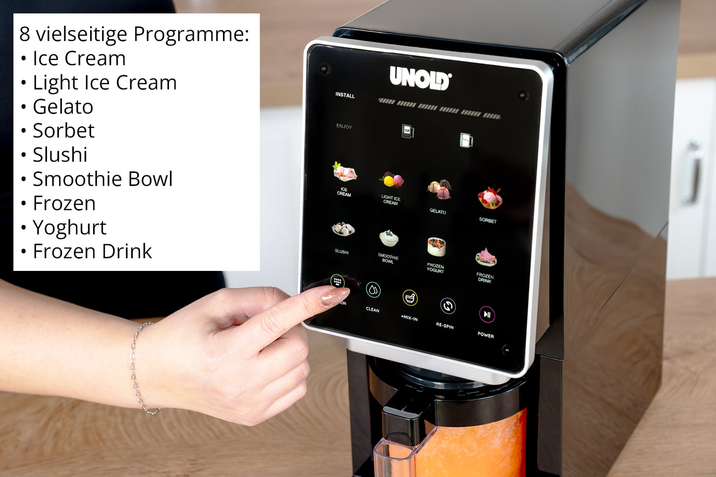 Eine Hand drückt einen Knopf an der schwarzen Eismaschine Cream Dream der Unold AG mit Digitalanzeige. Auf der linken Seite sind acht deutschsprachige Programme wie Eis, Sorbet und Joghurt aufgelistet. Im Inneren ist eine orangefarbene Mischung zu sehen.