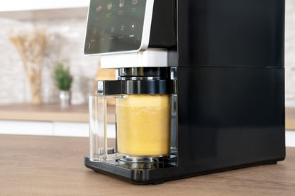 Eine Nahaufnahme der Unold AG Eismaschine Cream Dream, die auf einem Küchentisch steht und frischen gelben Saft in einen klaren Glasbehälter abfüllt, mit einem unscharfen Küchenhintergrund.