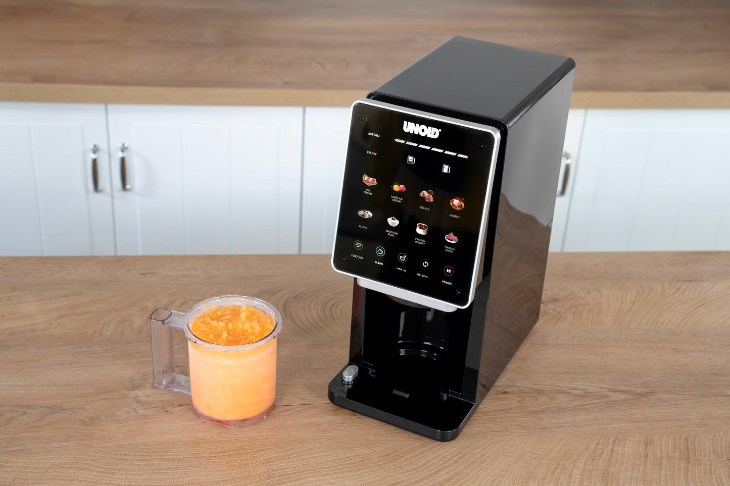 Die Unold AG Eismaschine Cream Dream steht auf einer hölzernen Arbeitsplatte neben einem klaren Becher mit einem Orangenmischgetränk. Im Hintergrund sind weiße Schränke und Holzarbeitsplatten zu sehen.