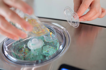 Eine Person gibt bunte Toppings in die mit mintgrünem Eis gefüllte Unold Eismaschine Cortina 1,5 L und hält mit der anderen Hand den durchsichtigen Plastikdeckel offen.