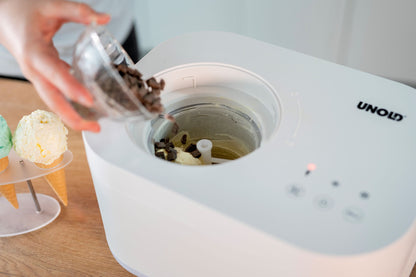 Eine Person füllt Schokoladenstückchen in eine mit Vanilleeis gefüllte Unold Eismaschine Snow 1 L. Daneben stehen drei Waffeln mit grünen und gelben Kugeln auf einem Teller. Die weiße Maschine trägt die Marke Unold.