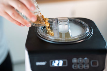 Eine Hand schüttet gehackte Nüsse aus einer kleinen Glasschale in den geöffneten Deckel der Unold Eismaschine Uno 1,2 L, deren Digitalanzeige 1:23 anzeigt.