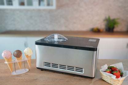 Die Unold Eismaschine Gusto 2 L steht auf einer Küchentheke neben drei Eistüten in einem Halter und einem Korb mit frischen Erdbeeren, im Hintergrund ist eine verschwommene Küchenkulisse zu sehen.