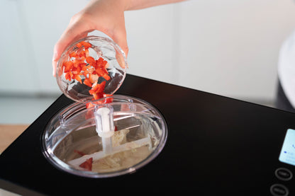 Eine Hand schüttet gehackte Erdbeeren aus einer kleinen Glasschale in die Unold Eismaschine Gusto 2 L, die teilweise gemischtes Eis enthält.