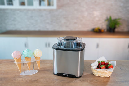 Die Eismaschine „Unold Eismaschine Gelato 1,5 L“ steht auf einer Küchentheke neben einem Halter mit drei Eistüten und einem Korb mit frischen Erdbeeren, im Hintergrund sind weiße Schränke und eine Rückwand zu sehen.