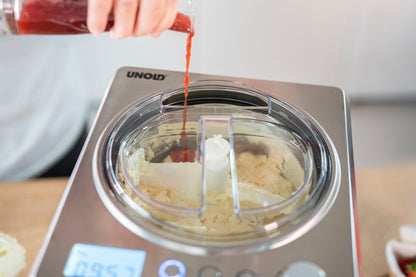 Jemand gießt rote Soße in eine Unold Eismaschine Exklusiv 2 L, die eine hellgelbe Eismischung enthält. Auf der Metalloberfläche der Maschine sind Display und Bedienelemente gut zu erkennen.