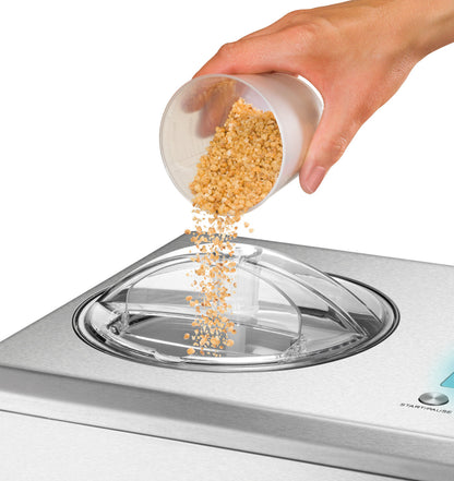 Eine Hand schüttet gehackte Nüsse aus einem Plastikbecher in den Zutatenschlitz der Unold Eismaschine Nobile (2,0 L) von Unold, einer Eismaschine aus Edelstahl, die mit einem Eisrezeptbuch geliefert wird.