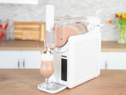 Die weiße Unold Slush-Eismaschine Susi gibt einen Schokoladenmilchshake mit Schlagsahne in ein hohes Glas mit gestreiftem Strohhalm und Löffel aus, das auf einer Küchentheke vor einem modernen Küchenhintergrund steht.