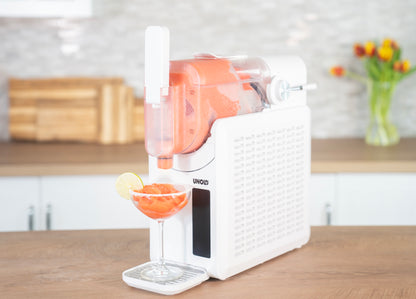 Die Unold Slush-Eismaschine Susi gibt ein leuchtend orangefarbenes Frozen Drink in ein Cocktailglas mit einer Zitronenscheibe aus, das in einer modernen, mit Blumen und Schneidebrettern geschmückten Küche steht.