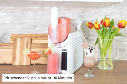 Die Unold Slush-Eismaschine Susi steht auf der Küchentheke und spritzt roten Slush in ein Glas. Daneben stehen zwei Getränke – ein rotes mit Limette, ein braunes mit Schlagsahne – sowie eine Vase mit Tulpen und Schneidebrettern im Hintergrund.