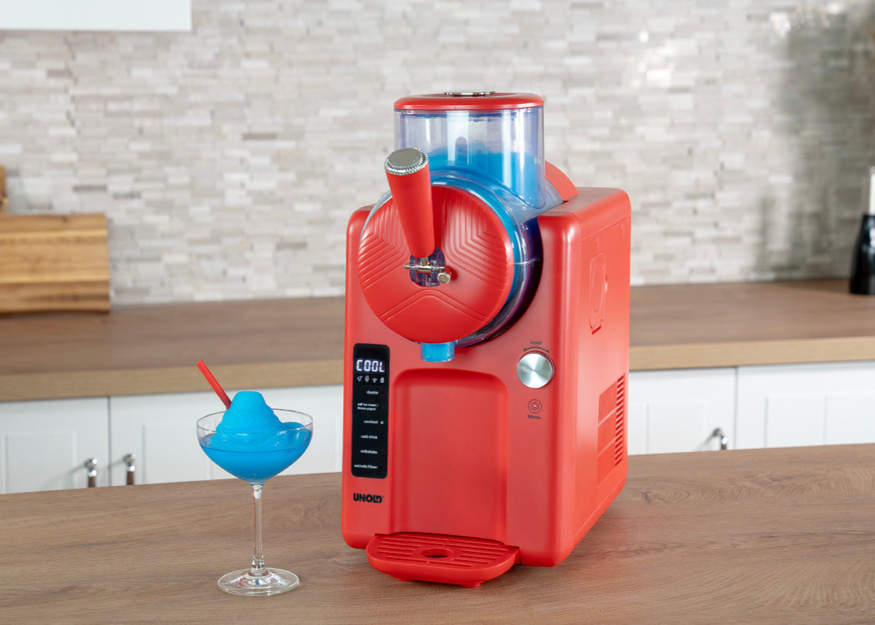 Die Unold 2in1 Slush- und Softeismaschine Sofie steht auf einer Küchentheke und spendet blauen Slush in ein Cocktailglas mit rotem Strohhalm. Dahinter befinden sich eine geflieste Aufkantung und ein Holzdekor.