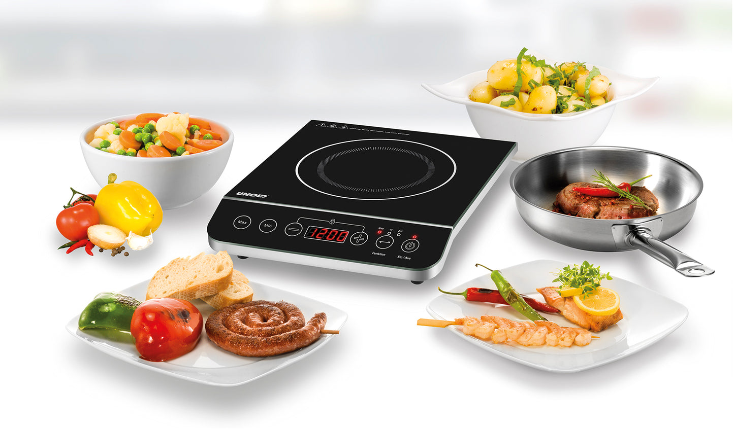 Die Unold AG Einzel-Induktionskochplatte - Schwarz/Edelstahl 581051 (B-Ware) ist umgeben von Tellern und Schalen mit gekochtem Gemüse, gegrilltem Fleisch, Brot, Wurst und einer Pfanne mit Steak und Kartoffeln.
