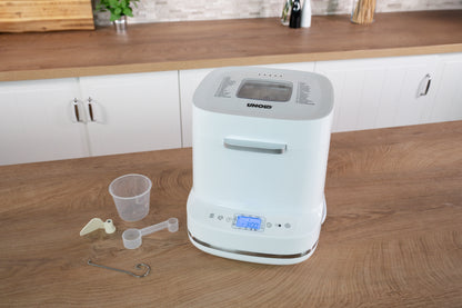 Der Weißbrotbackautomat der Unold AG Backmeister Bernd steht auf einer Holztheke mit Messbecher, Löffel, Haken und Knetmesser im Vordergrund. Auf dem Display steht 3:00, im Hintergrund sind weiße Schränke zu sehen.