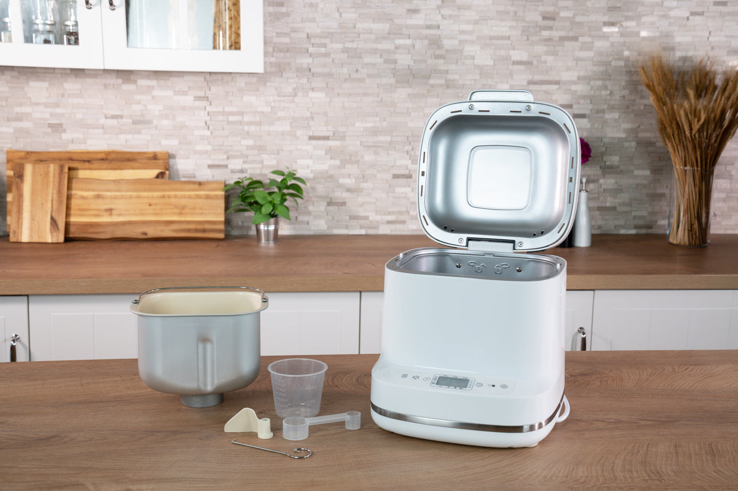 Der Unold AG Backmeister Bernd Brotbackautomat steht offen auf einer modernen Küchentheke, zusammen mit der Brotbackform, dem Messbecher und dem Löffel, dem Knetmesser und dem Haken. Die stilvolle Umgebung besteht aus Schränken, Pflanzen und Schneidebrettern.