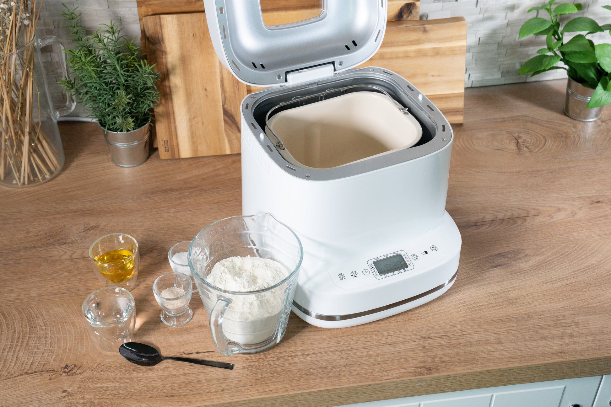 Der Brotbackautomat der Unold AG Backmeister Bernd steht mit geöffnetem Deckel auf einer hölzernen Arbeitsplatte, Mehl, Wasser, Öl, Salz und Hefe sind in Glasbehältern abgemessen. Ein schwarzer Löffel liegt daneben, im Hintergrund stehen Grünpflanzen und Schneidebretter.