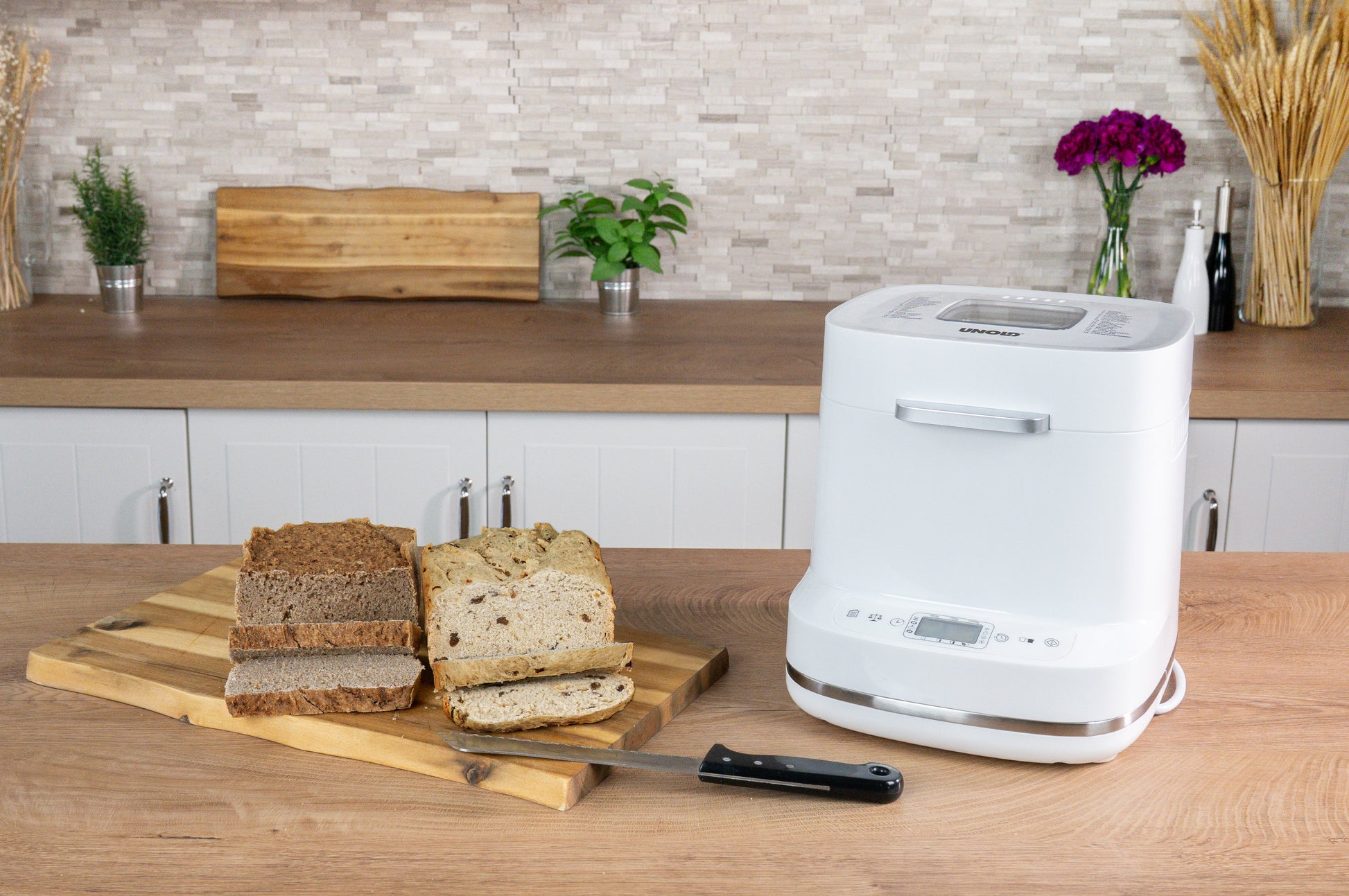 Der Brotbackautomat der Unold AG Backmeister Bernd steht auf einem Küchentisch neben einem hölzernen Schneidebrett mit verschiedenen Brotscheiben und einem Brotmesser; im Hintergrund sind Pflanzen, Blumen und ein weiteres Schneidebrett zu sehen.