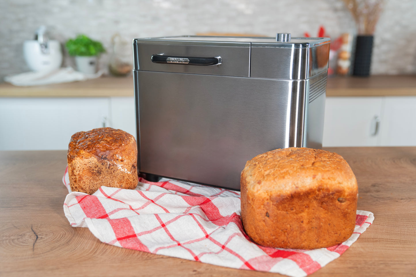 Der Unold Brotbackautomat BackProfi Nussspender der Unold AG steht auf einer Holztheke mit zwei Broten auf einem rot-weiß karierten Tuch in einer hellen Küchenumgebung.
