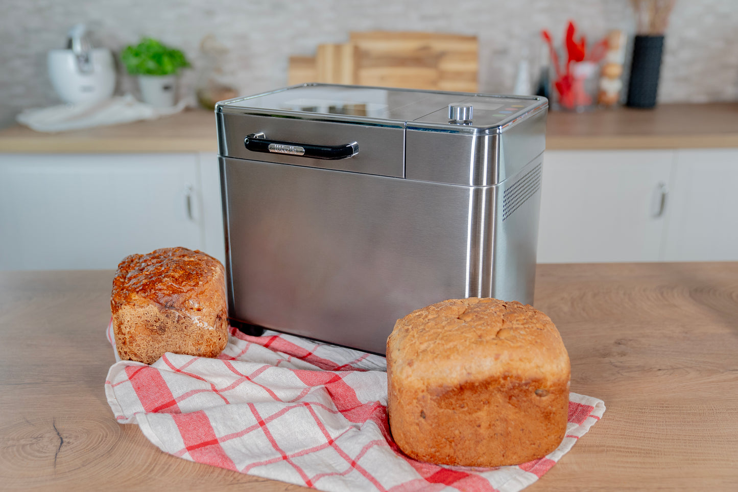 Der Unold AG Brotbackautomat BackProfi mit Nussspender und Rezeptheft steht auf einer Küchentheke neben zwei frischen Broten auf einem rot-weiß-karierten Tuch, im Hintergrund eine sanft verschwommene Küche.