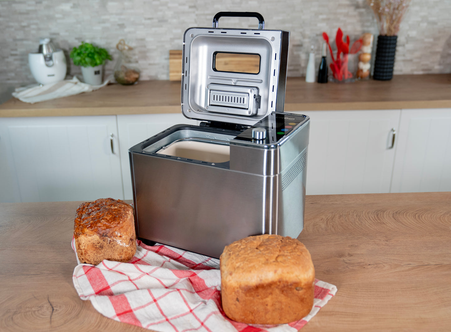 Der Unold AG Brotbackautomat BackProfi Nussspender (mit Rezeptheft) steht offen auf einer Küchentheke neben zwei Broten und einem rot-weiß-karierten Tuch, im Hintergrund sind Küchengeräte zu sehen.