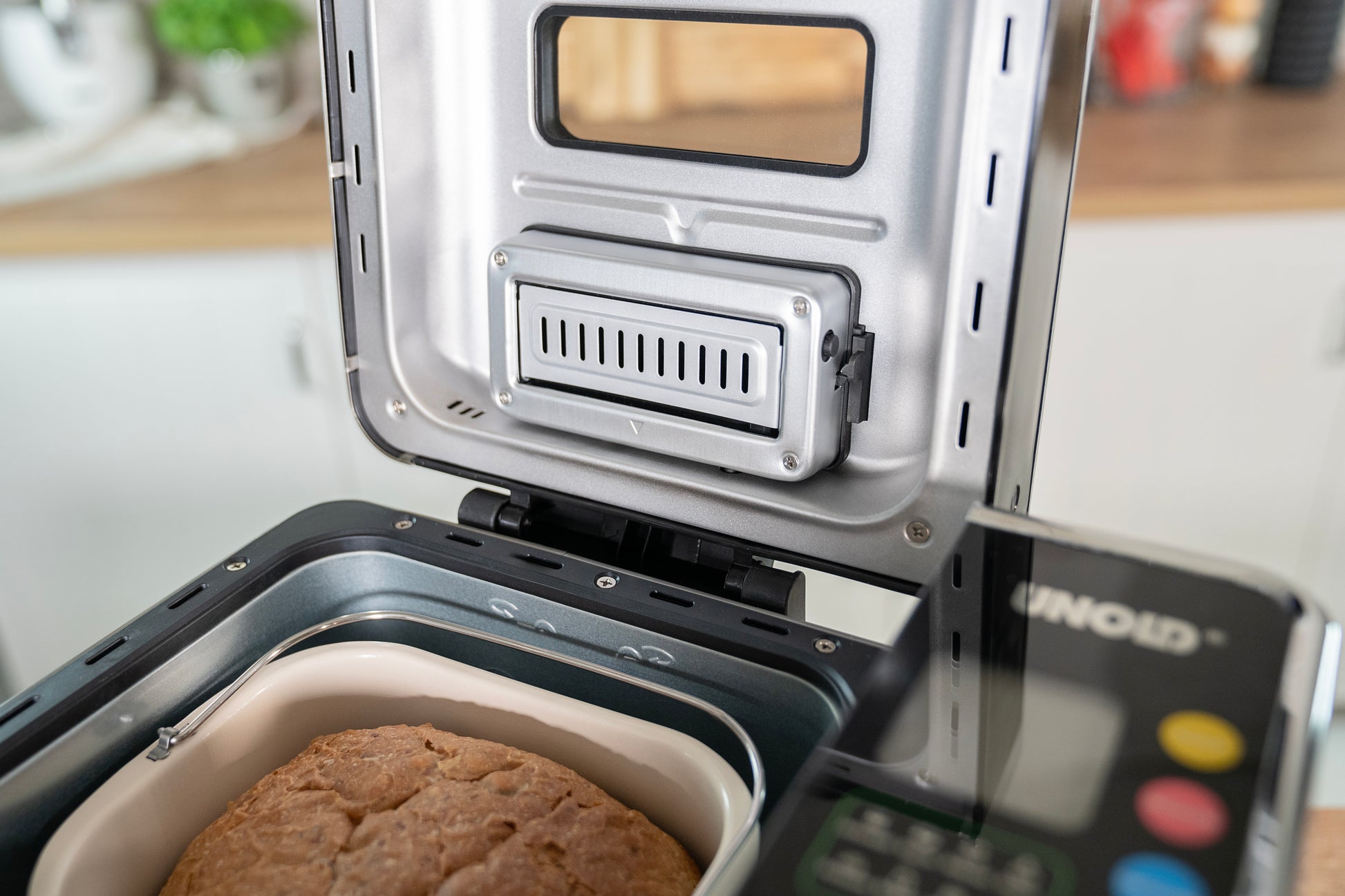 Ein Brot wird im Brotbackautomat BackProfi Nussspender inkl. Rezeptheft der Unold AG gebacken. Rezeptheft Brotbackautomat, im Hintergrund sind das Bedienfeld und ein Teil der Küche zu sehen.