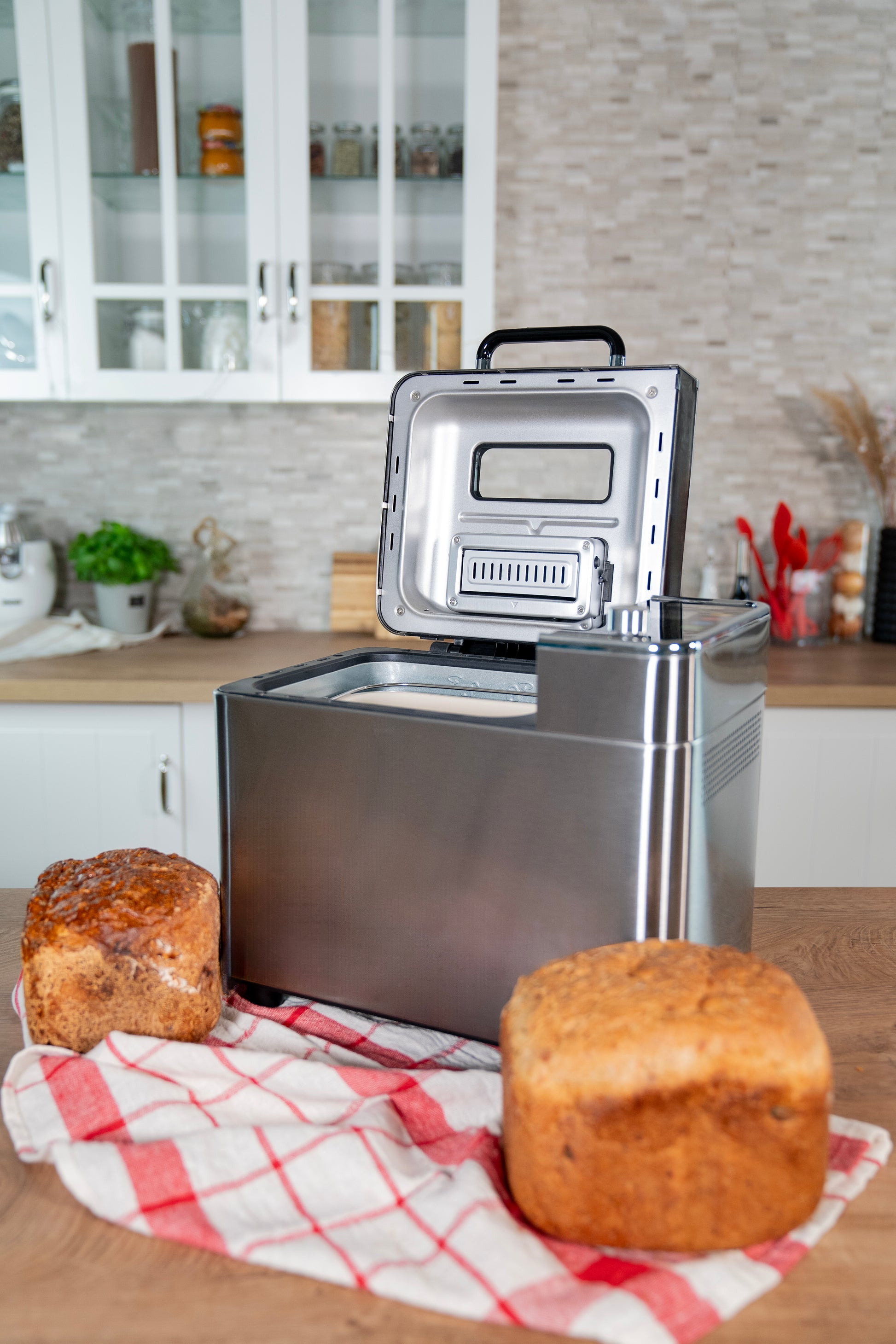 Der Unold AG Brotbackautomat BackProfi Nussspender steht mit geöffnetem Deckel auf einer Küchentheke neben zwei Broten auf einem rot-weiß-karierten Tuch, im Hintergrund sind Schränke und Utensilien zu sehen.