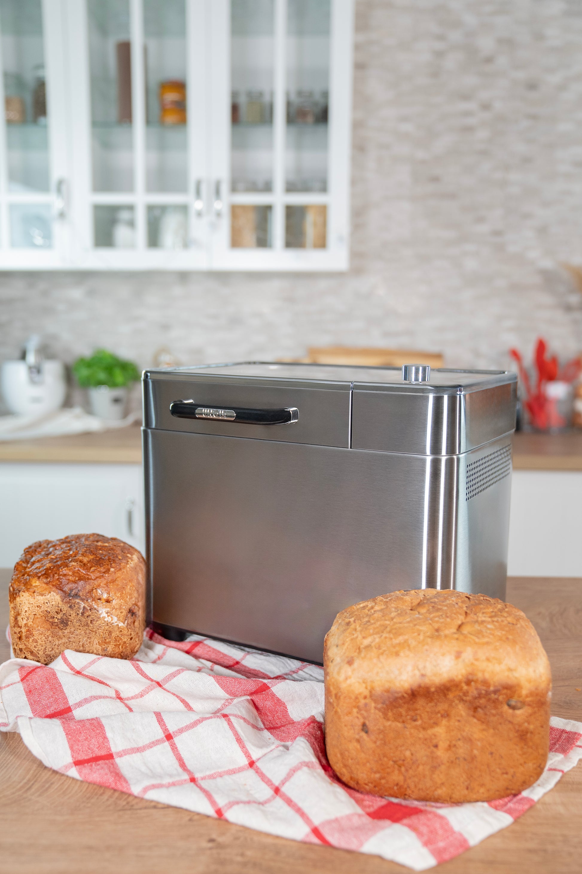 Der Unold AG Unold Brotbackautomat BackProfi Nussspender inkl.. Rezeptheft sitzt auf einer Küchentheke mit zwei Broten auf einem rot-weiß-karierten Tuch; im Hintergrund sind Schränke und Utensilien zu sehen.
