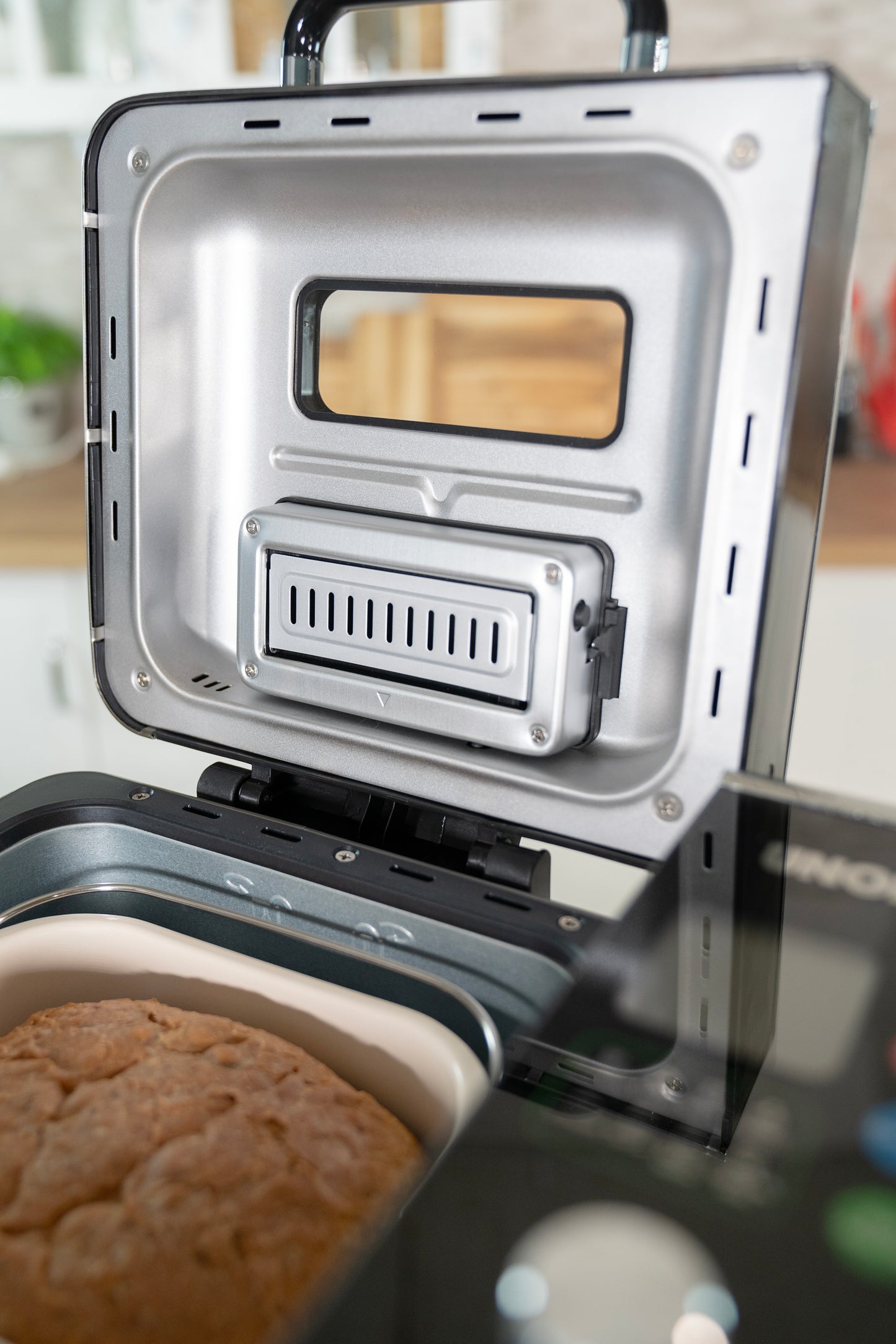 Der Unold AG Brotbackautomat BackProfi Nussspender mit geöffnetem Metalldeckel und Lüftungsschlitz offenbart ein frisch gebackenes Brot im Inneren. Der unscharfe Küchenhintergrund hebt das mitgelieferte Rezeptheft der Unold AG hervor.